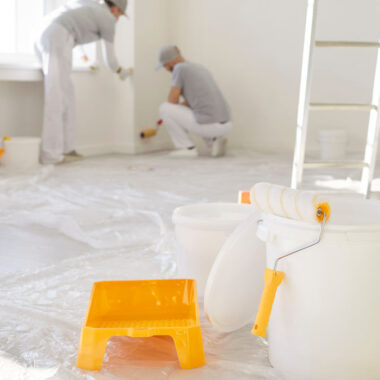 Professional painters working indoors with rollers, paint buckets, and protective floor covering during a home interior painting project.