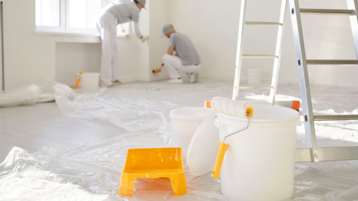 Professional painters working indoors with rollers, paint buckets, and protective floor covering during a home interior painting project.